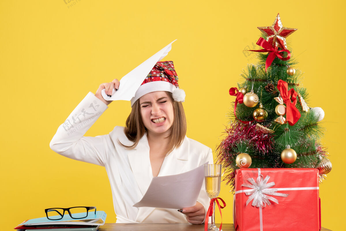 快乐正面图：女医生坐在黄色的桌子上 手里拿着圣诞礼物 手里拿着文件坐着女性女医生