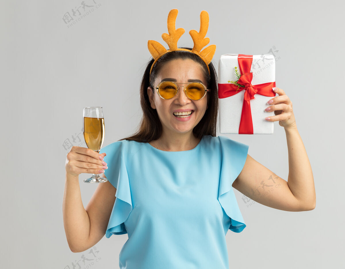鹿身着蓝色上衣的快乐年轻女子 戴着滑稽的鹿角边和黄色眼镜 手里拿着一杯香槟和圣诞礼物 笑容满面蓝色香槟礼物