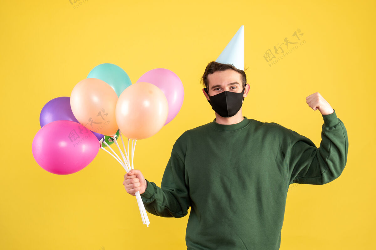 幸福正面图：戴着派对帽的年轻人 五颜六色的气球展示着黄色的肌肉年轻喜剧演员人