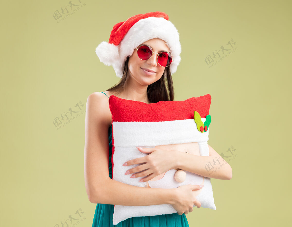 快乐戴着太阳眼镜 戴着圣诞帽 抱着圣诞枕头的年轻高加索女孩高兴极了空间新的年轻