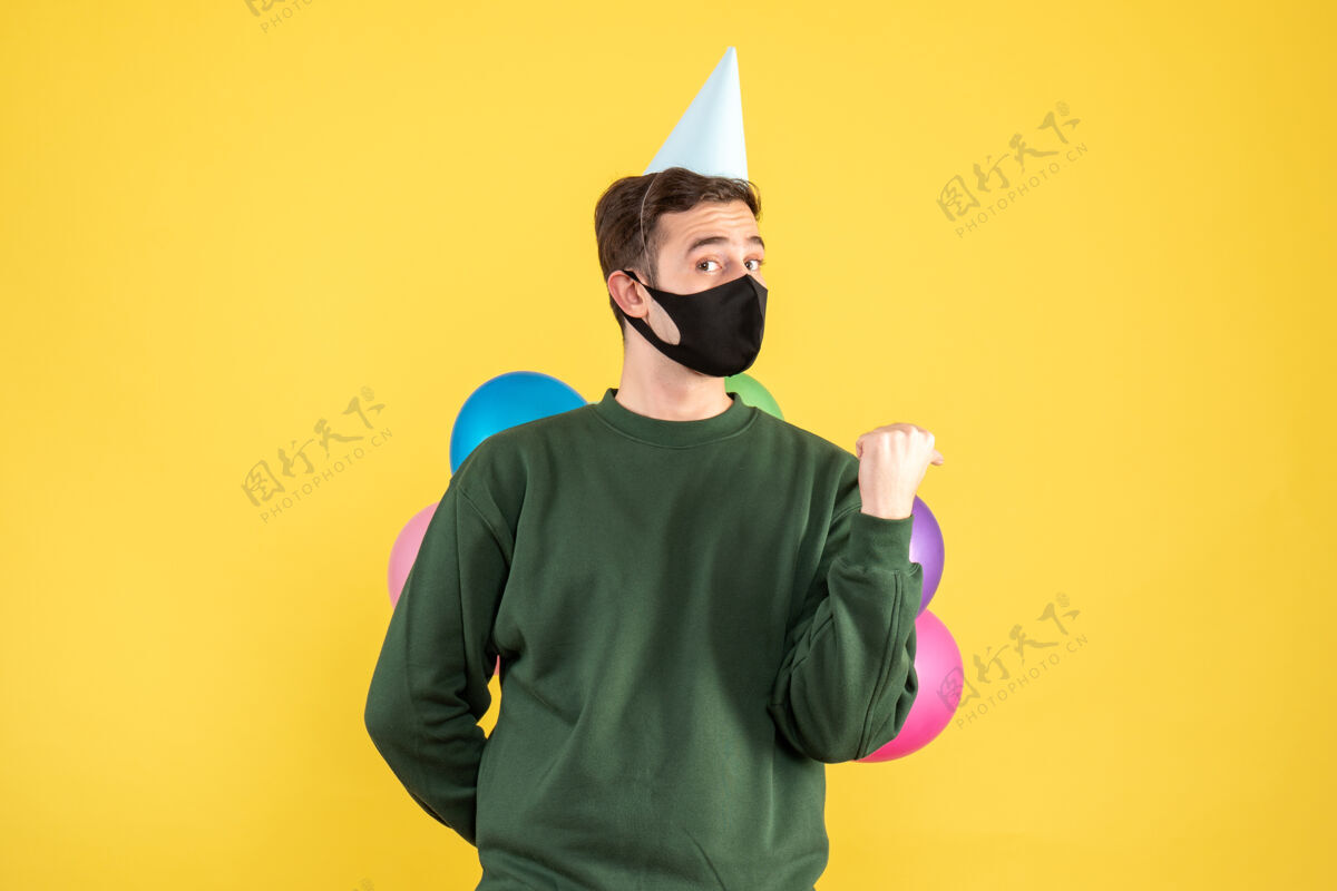 保护正面图：戴着派对帽 站在黄色地面上的彩色气球的年轻人年轻人面具掩护