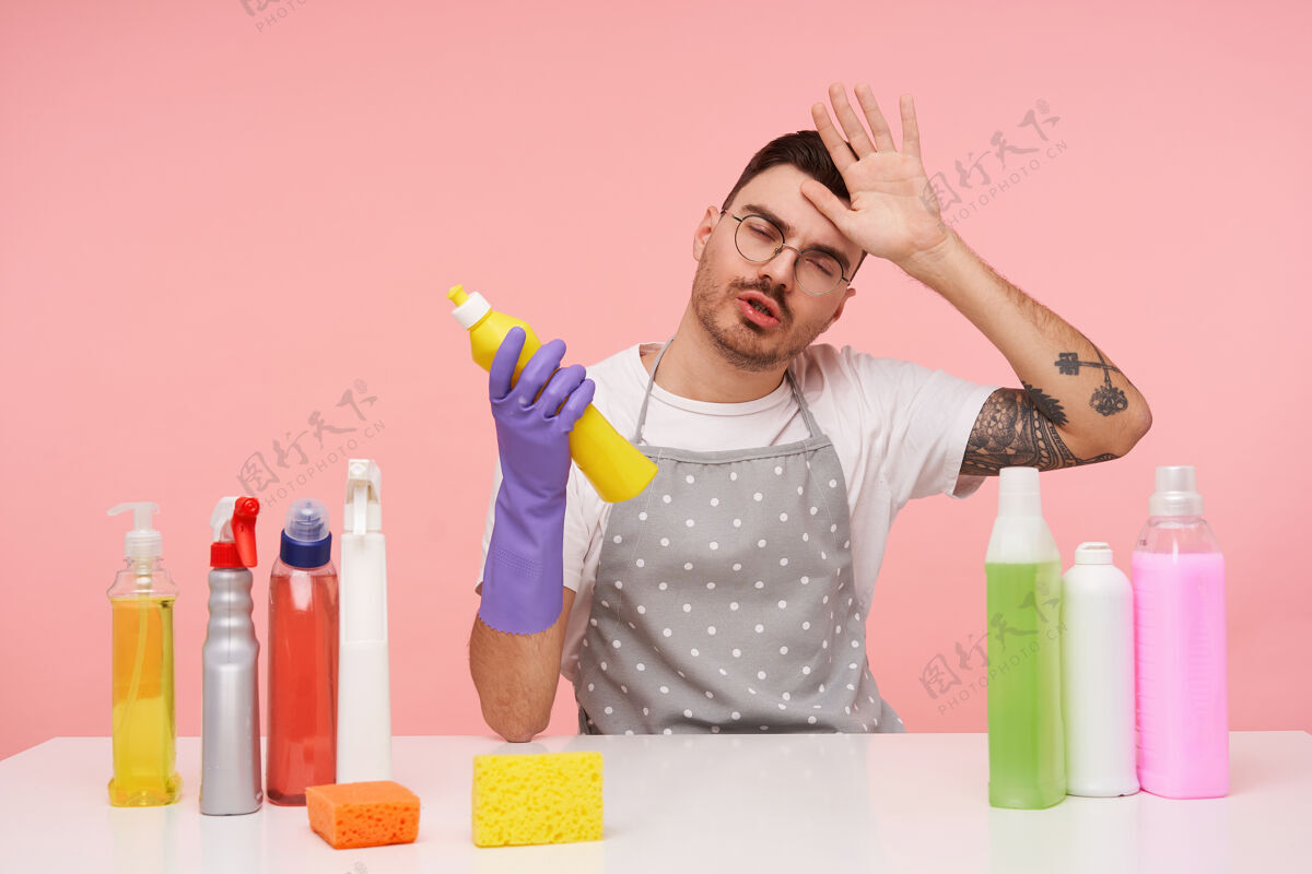 精疲力尽年轻的棕色头发的男性纹身画像举起疲惫的手到他的脸上 而坐在粉红色的洗涤剂 与清洁室休息深色胡须抬高