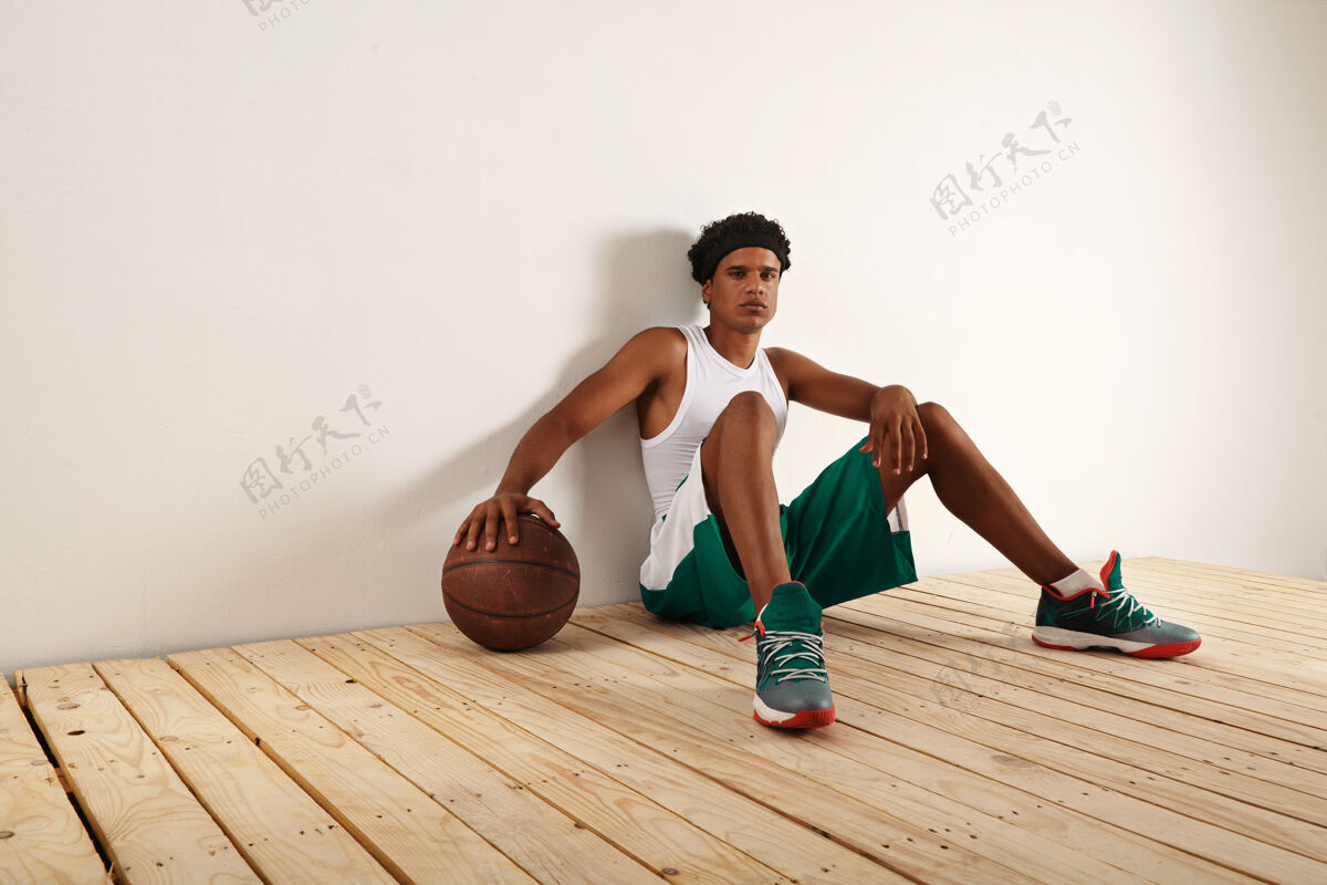 Smart穿着绿色和白色篮球服的疲惫而体贴的黑人篮球运动员坐在浅色的木地板上 把手放在一个肮脏的棕色篮球上BrownGymSport