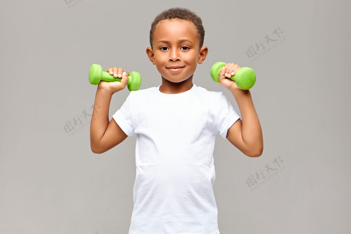 童年快乐的非洲裔美国男孩的肖像 瘦瘦的胳膊 微笑着快乐的同时 在健身房锻炼两个哑铃 要建立强大的健康的运动身体健身和儿童设备体重运动