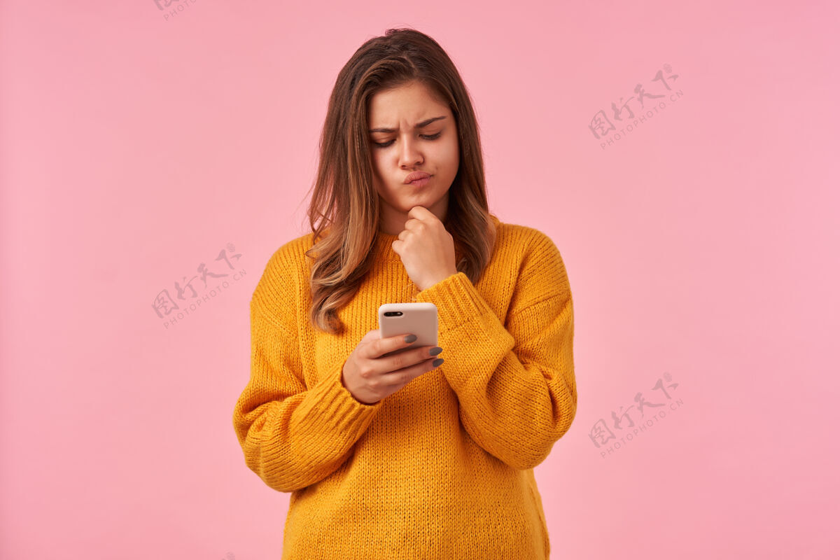 温暖困惑的年轻漂亮的棕色头发的女性扭曲她的嘴和皱眉困惑地看着她的手机屏幕眉毛 孤立的粉红色智能手机自然色深色