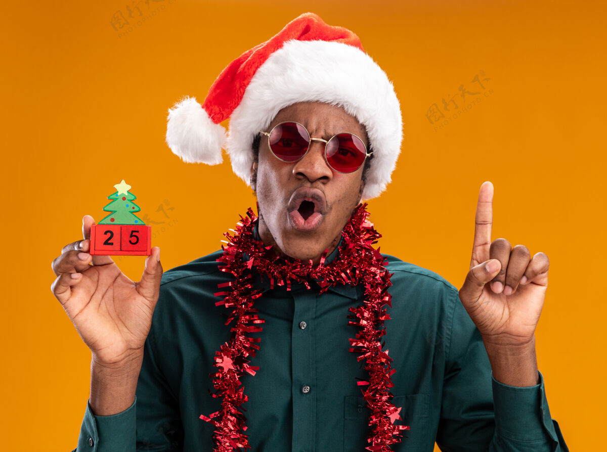 圣诞节戴着圣诞帽 戴着花环 戴着太阳镜 拿着玩具方块的非洲裔美国人 25岁的他看起来很惊讶 食指站在橙色的墙上站立显示男人