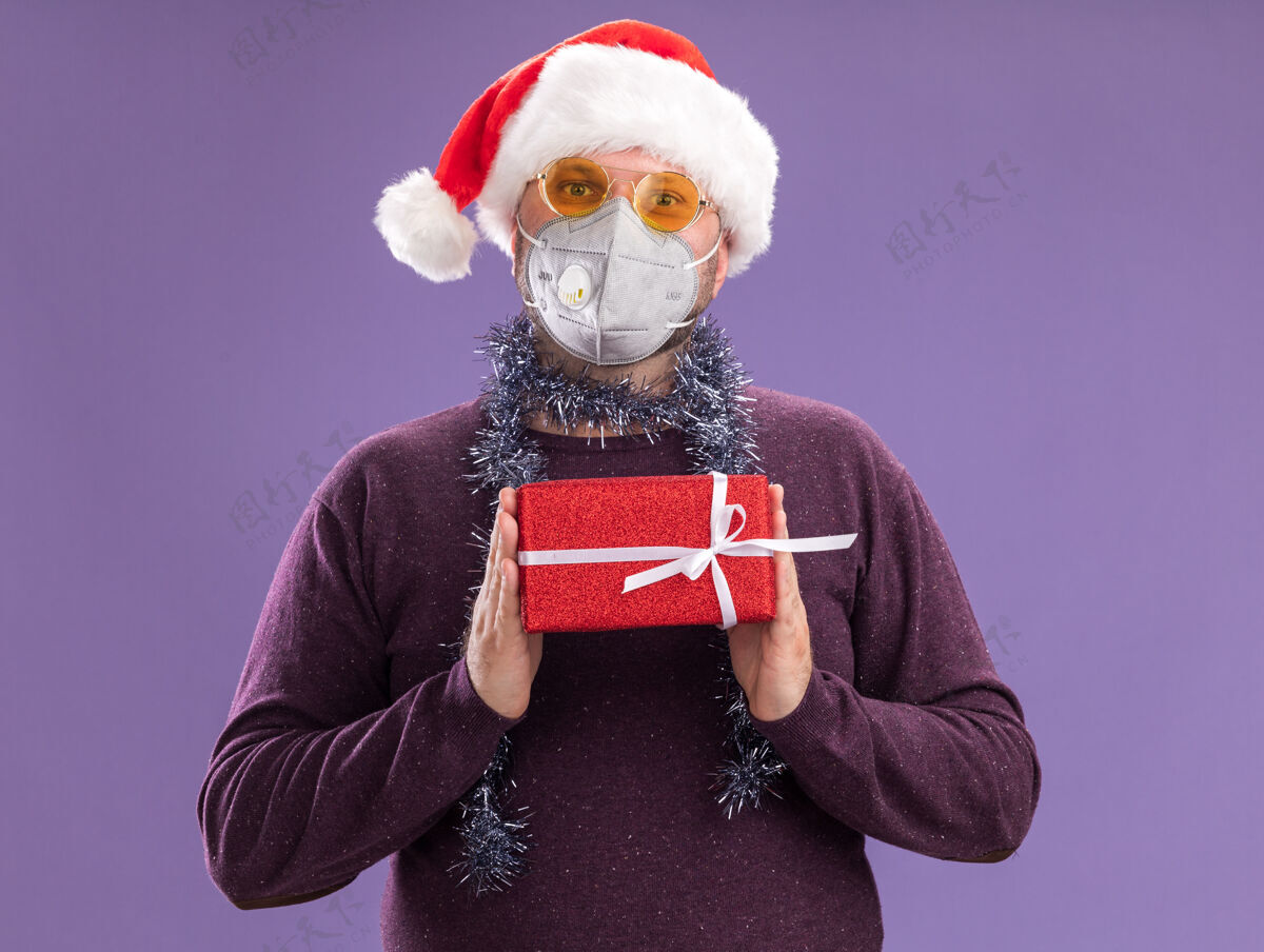 金属丝中年男子戴着圣诞帽 戴着防护面具 脖子上戴着金属丝花环 戴着眼镜 手里拿着礼包 隔离在紫色的墙上包装保护中年