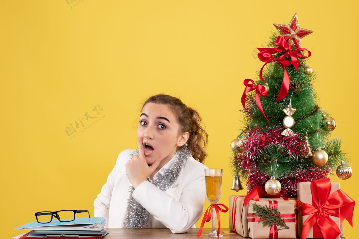 女性正面图女医生围坐在黄色背景上的圣诞礼物和圣诞树旁成人女医生人