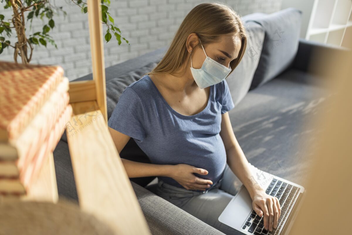 职业孕妇戴着医用面罩坐在沙发上用笔记本电脑工作母亲水平工作