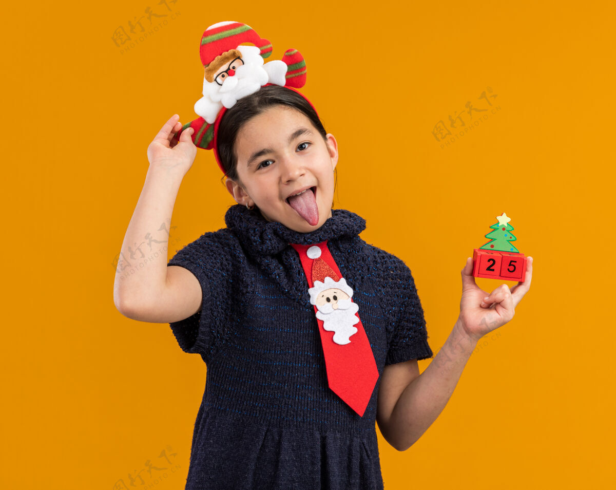 小快乐的小女孩穿着针织连衣裙 头上系着一条红色领带 带着有趣的边 手里拿着一个玩具立方体 嘴里还伸着圣诞节的日期戴着领带方块