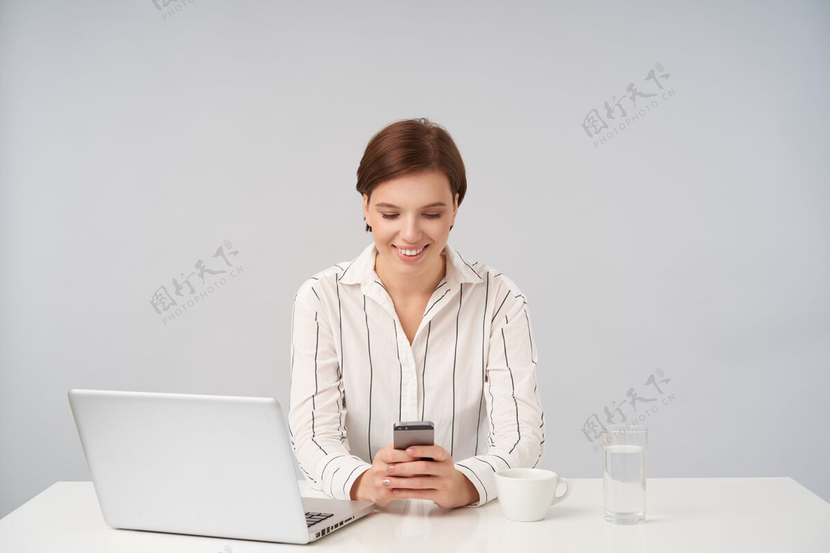 积极可爱的棕色头发的年轻漂亮女人留着时髦的短发 手里拿着智能手机 和朋友聊天时面带微笑 孤独地站在白色的地板上短黑发办公室