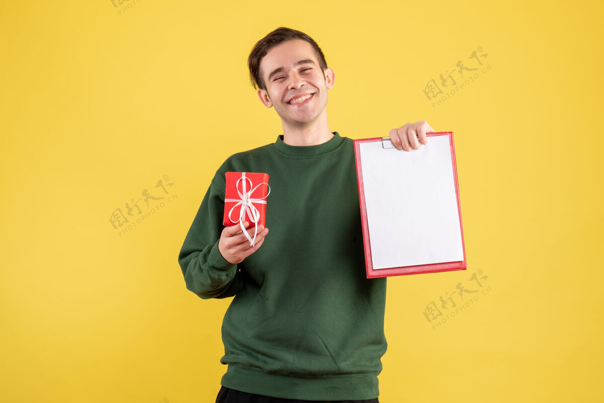 站立正面图：穿着绿色毛衣的年轻人拿着礼物 剪贴板站在黄色的地板上毛衣地方年轻人