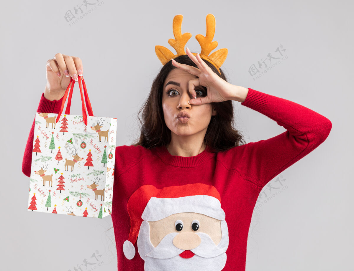 驯鹿印象深刻的年轻漂亮女孩穿着驯鹿鹿角头带和圣诞老人毛衣拿着圣诞礼物袋看着相机做的样子手势孤立在白色背景上拿着头带漂亮