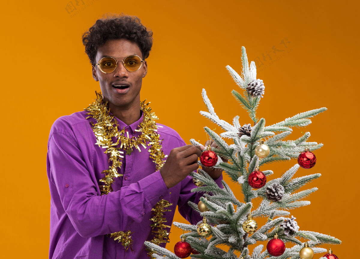 花环令人印象深刻的年轻非裔美国人戴着眼镜 脖子上戴着金箔花环 站在橙色背景的装饰圣诞树旁圣诞饰品装饰站着