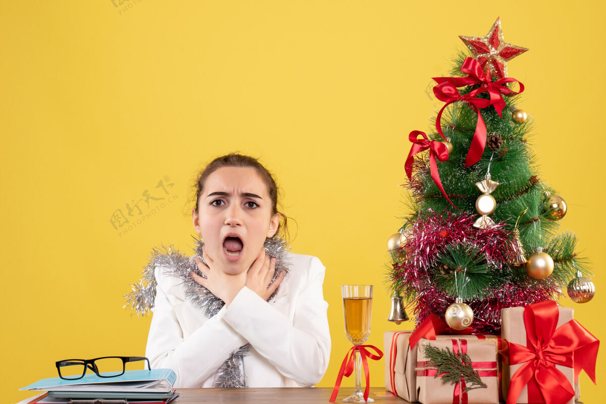 喉咙正面图：坐在桌子后面的女医生喉咙有问题 背景是黄色的圣诞树和礼品盒电脑护士桌子