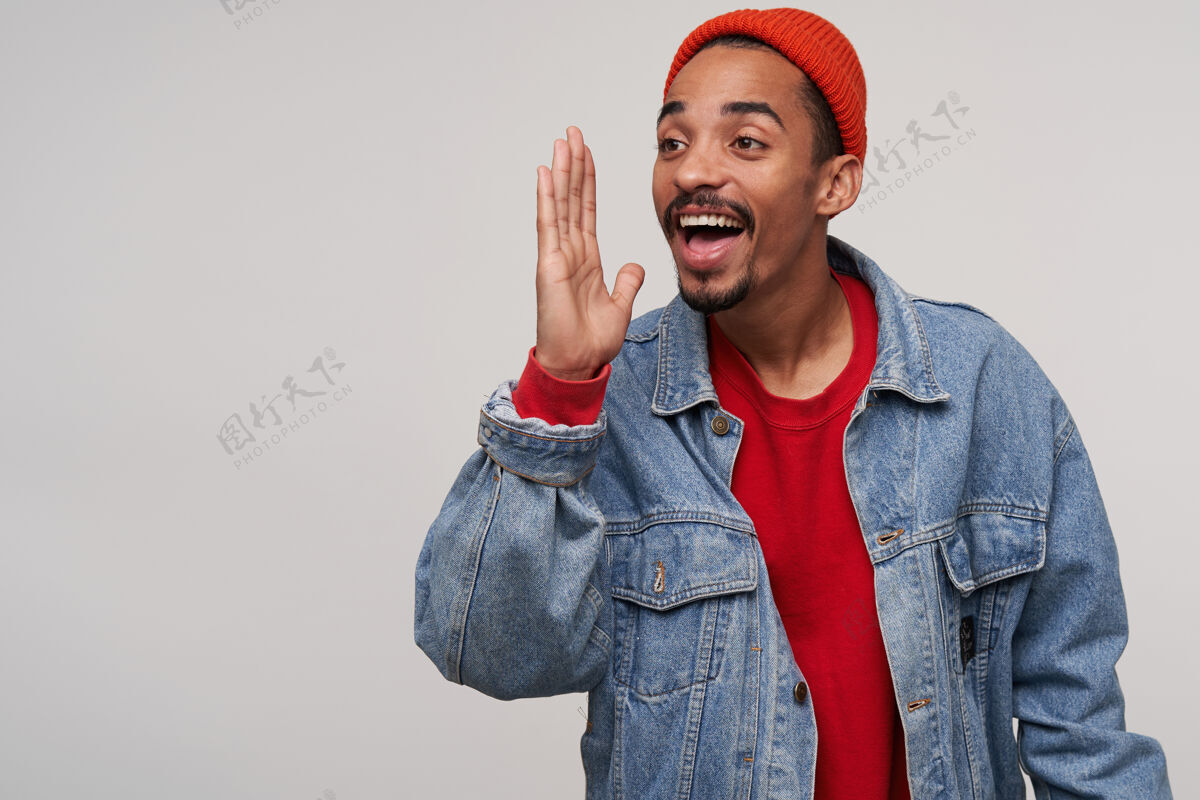 非洲人室内写真：一个快乐可爱的年轻人 留着胡子 皮肤黝黑 戴着红帽子 穿着红色套头衫和牛仔裤 在白色的床上摆着姿势 手掌高举 笑容满面男性站着不留胡子