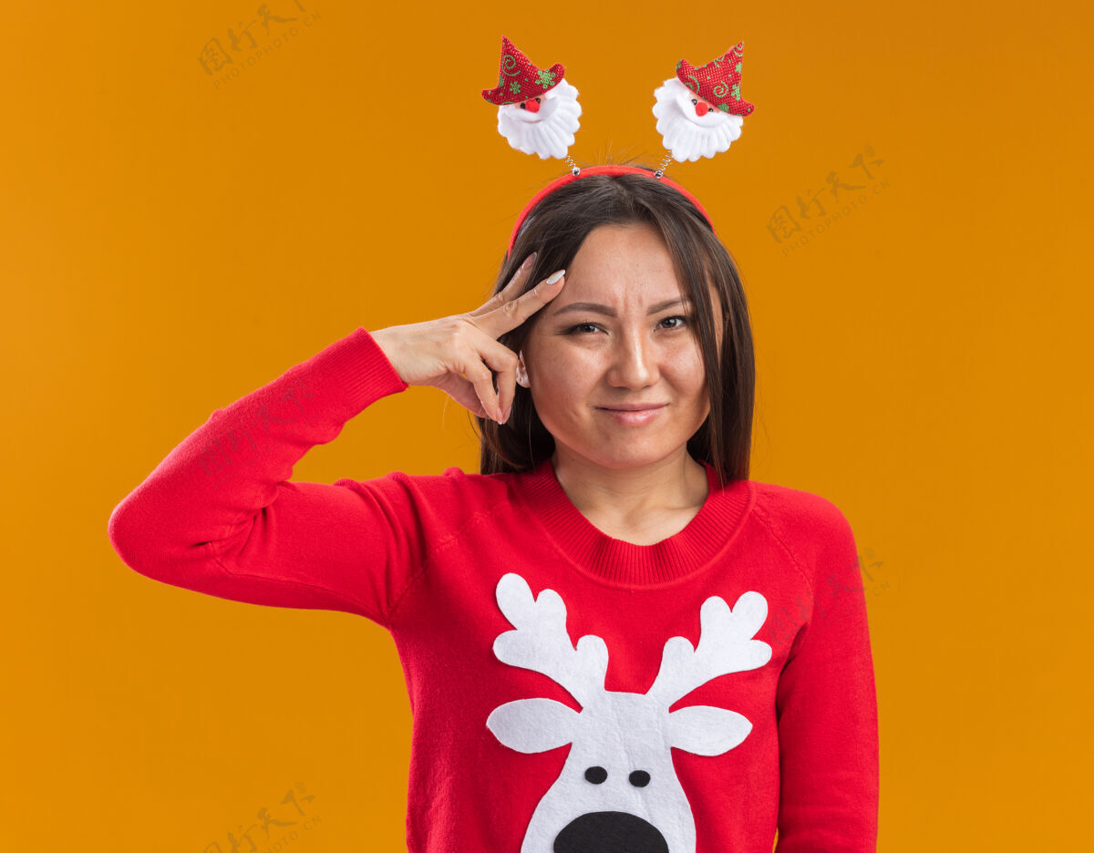 圣诞节紧张的年轻亚洲女孩戴着圣诞发箍和毛衣把手指放在隔离在橙色墙上的头上手指紧张放