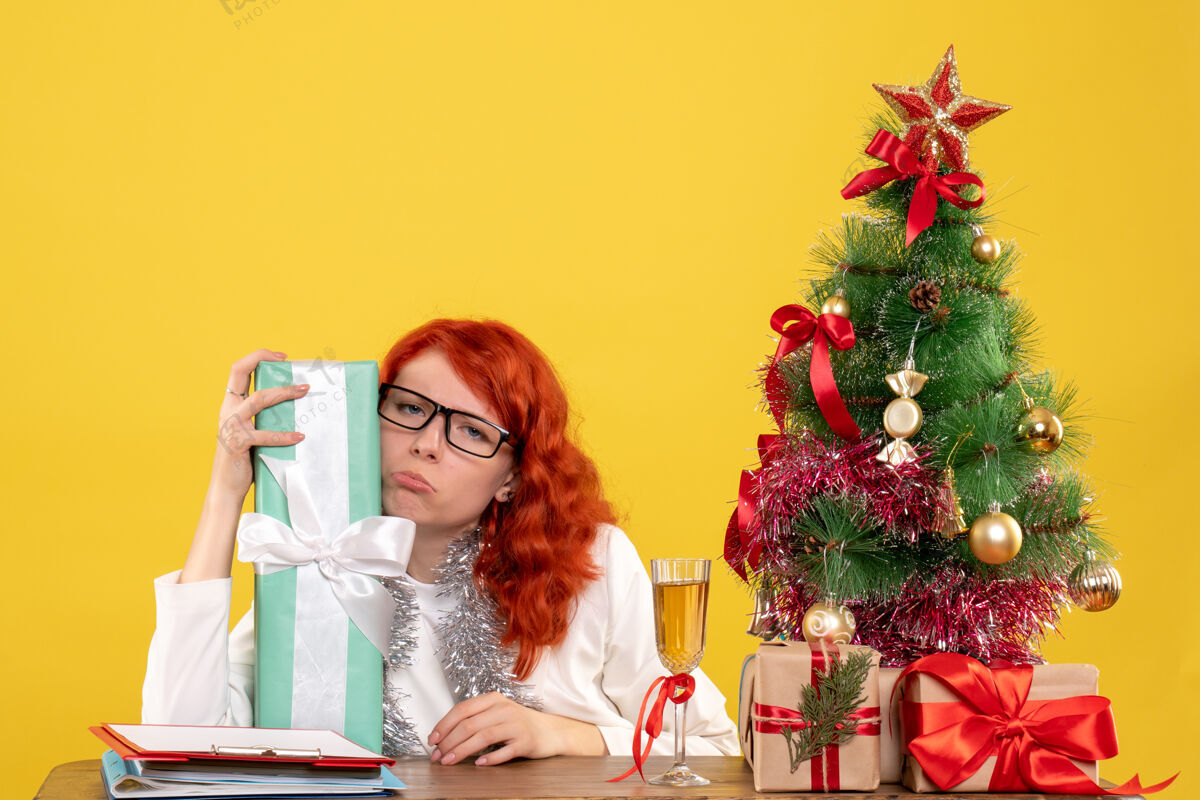 花前视图：女医生拿着圣诞礼物和黄色背景上的圣诞树坐着圣诞节坐着医生