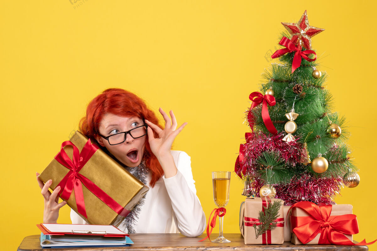 前面前视图：女医生拿着圣诞礼物和黄色背景上的圣诞树坐着生活人快乐