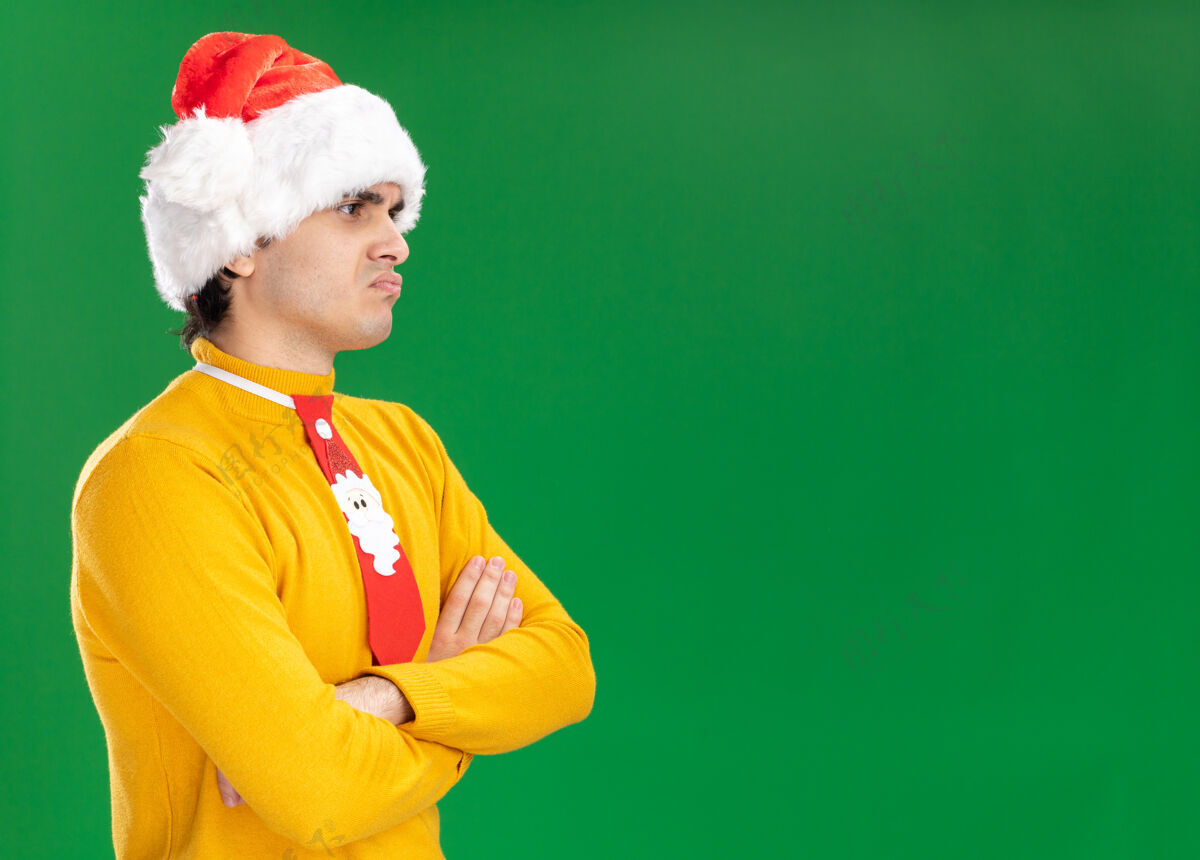 圣诞老人愤愤不平的年轻人穿着黄色高领毛衣 戴着圣诞老人帽 打着滑稽的领带 不高兴地交叉双臂站在绿色的背景上 向一旁望去旁边领带交叉