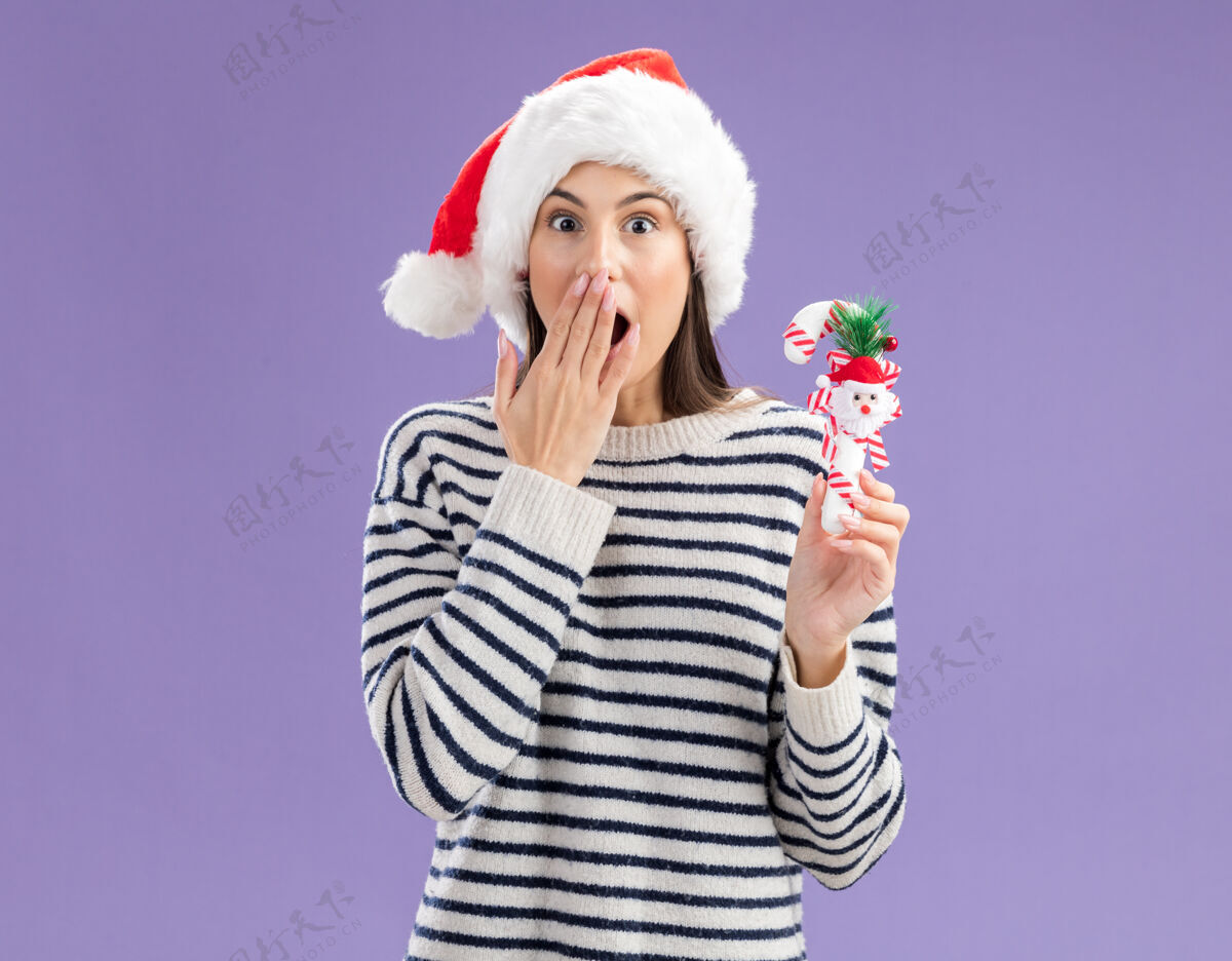 新戴着圣诞帽的年轻白人女孩惊讶地把手放在嘴上 手里拿着糖果棒圣诞老人年轻圣诞快乐