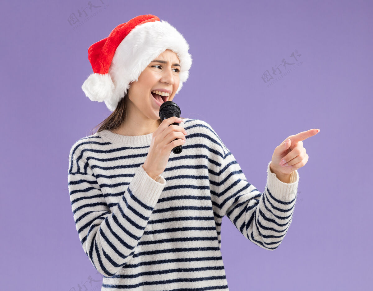 假装快乐的年轻白人女孩戴着圣诞帽拿着麦克风假装唱歌看着在紫色背景上与复制空间隔离的一面圣诞老人新年年轻