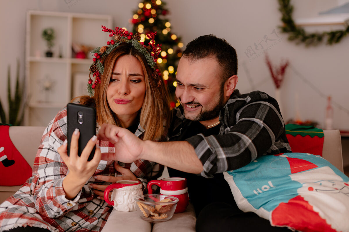 圣诞快乐一对幸福的年轻夫妇坐在沙发上喝着茶 在装饰好的房间里玩着智能手机 背景是圣诞树杯子年轻快乐