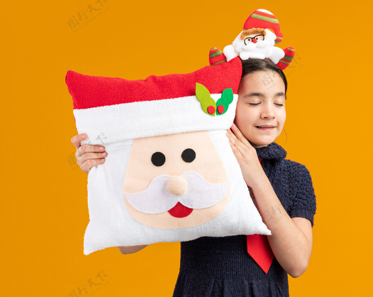 领带穿着针织连衣裙的快乐小女孩戴着红色领带 头上戴着滑稽的边 抱着圣诞枕头 闭着眼睛微笑着快乐微笑年