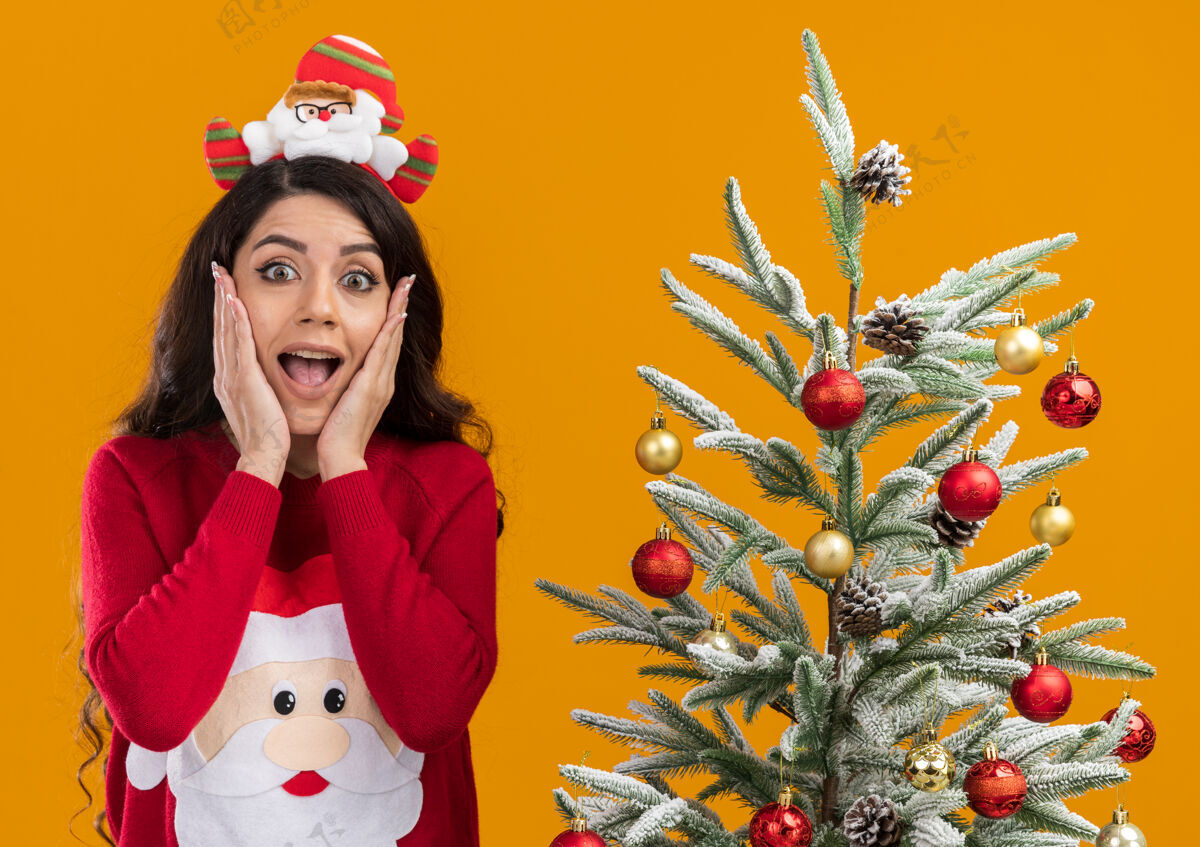 近兴奋的年轻漂亮女孩戴着圣诞老人的头带和毛衣站在装饰过的圣诞树旁 手放在脸上看着橙色背景上的相机脸兴奋橙色