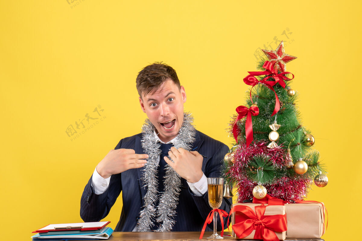 树前视图：商人指着自己坐在圣诞树旁的桌子 展示黄色的礼物桌子新郎生意人
