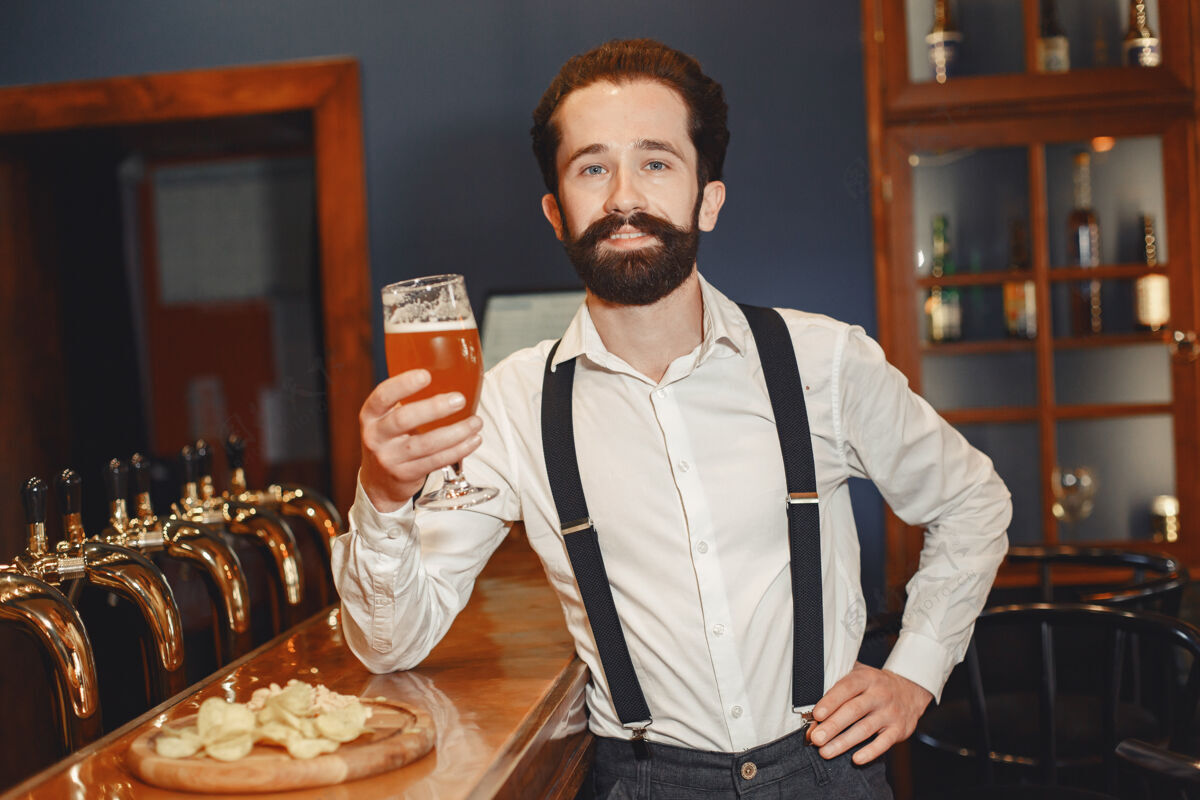 头发一个留着胡子的男人站在吧台旁 喝着酒杯里的酒啤酒成人男性