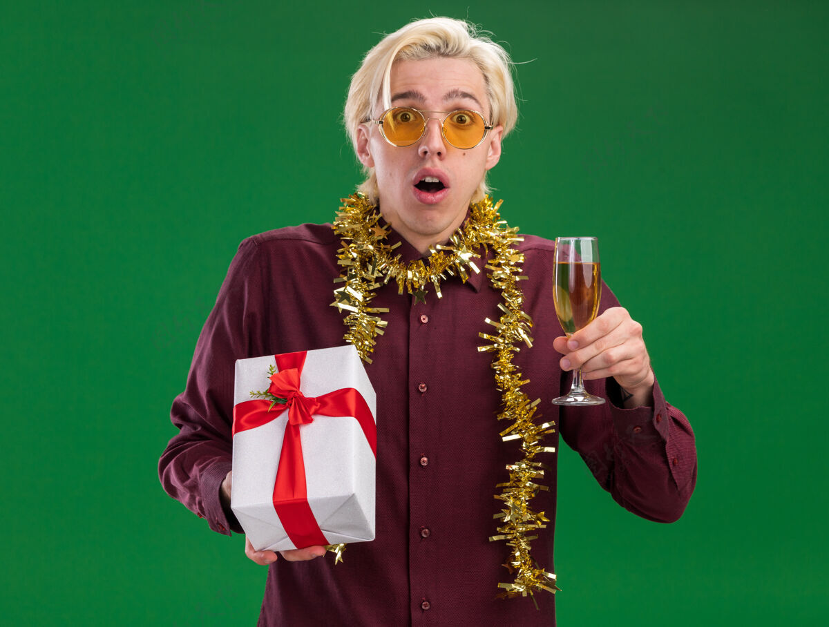 镀金令人印象深刻的年轻金发男子戴着一副眼镜 脖子上戴着金属丝花环 手里拿着一杯香槟和一个孤立在绿色墙上的礼包圣诞节金发脖子