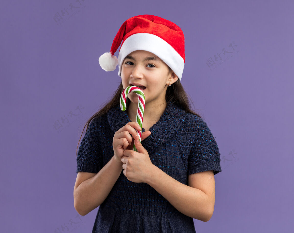 快乐快乐快乐的小女孩穿着针织连衣裙戴着圣诞帽拿着糖果手杖脸上带着微笑要咬手杖帽子糖果新年