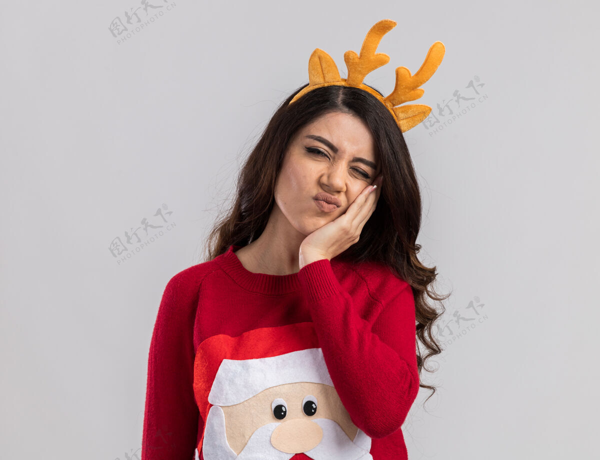 漂亮疼痛的年轻漂亮女孩戴着驯鹿鹿角头带和圣诞老人毛衣手放在脸上牙疼闭着一只眼睛看封闭的保管圣诞老人