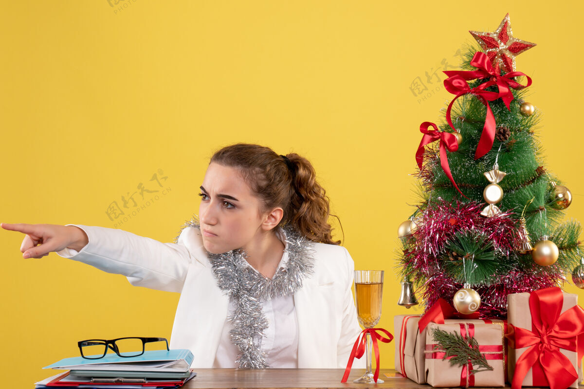 女医生正面图：女医生坐在桌子后面 指着黄色背景上的圣诞树和礼品盒人圣诞节快乐
