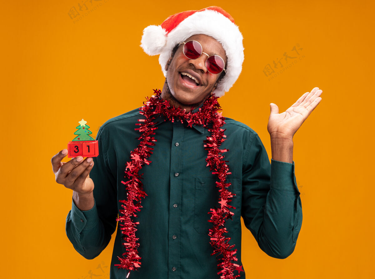 非洲非洲裔美国人戴着圣诞帽 戴着花环 戴着太阳镜 举着玩具立方体和新年约会 快乐快乐地举起手臂站在橙色的墙上圣诞老人男人圣诞节