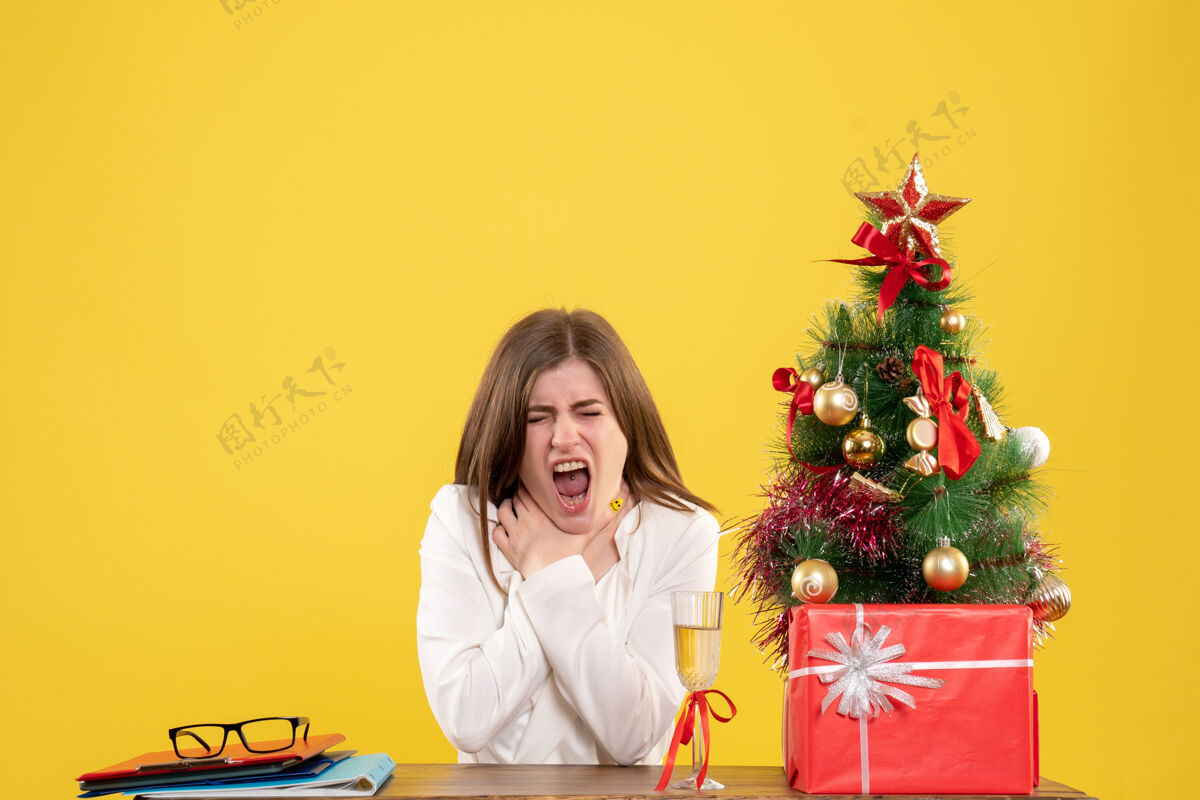 医生正面图：女医生坐在桌子前 喉咙痛 背景是黄色的圣诞树和礼品盒健康圣诞节情感