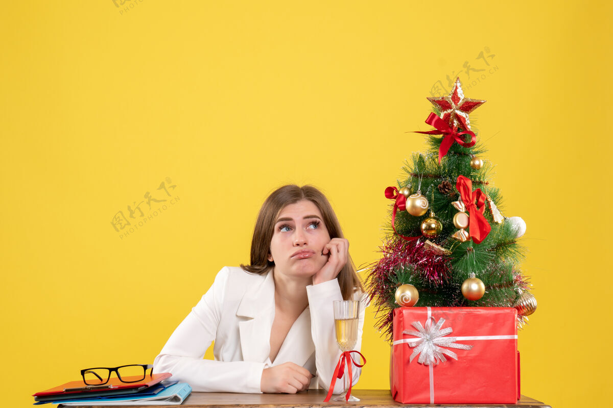 快乐正面图：女医生坐在黄色背景的桌子前 放着圣诞树和礼品盒女性医院微笑