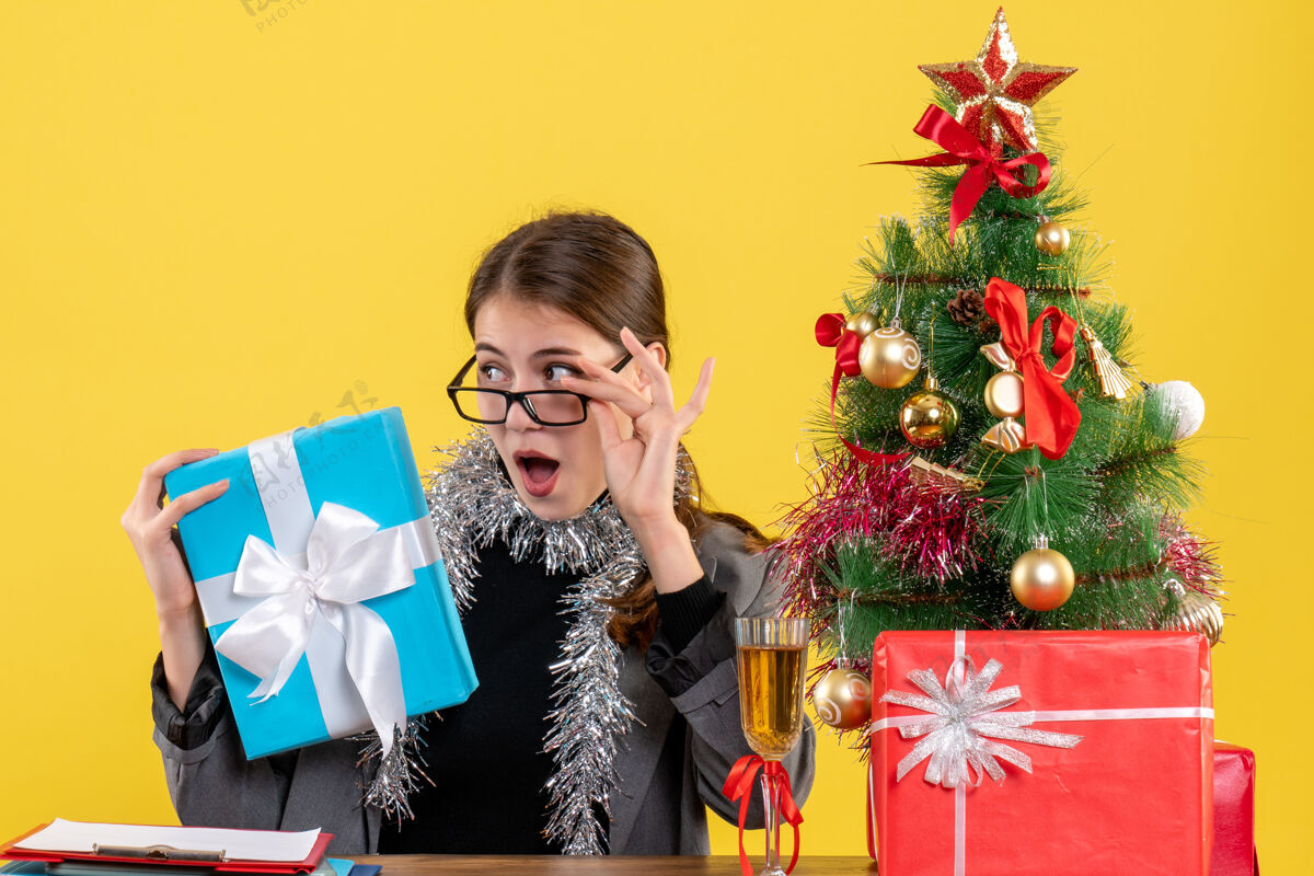 圣诞节正面图戴眼镜的大眼睛女孩坐在桌边摘下眼镜圣诞树和鸡尾酒礼物眼镜坐着树