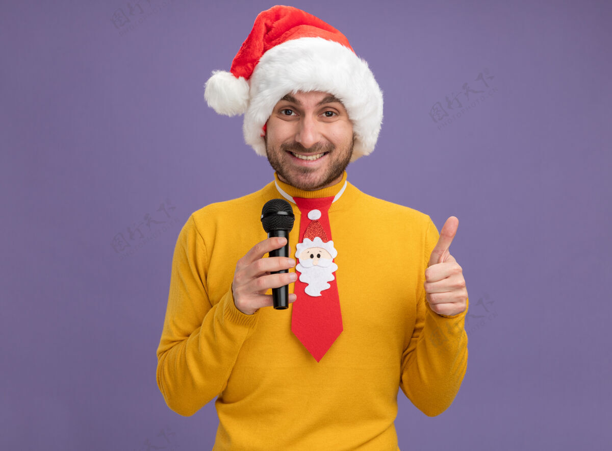 展示快乐的白人年轻人戴着圣诞帽 打着领带 拿着麦克风 在紫色的墙上孤立地竖起大拇指 留着复印空间拇指圣诞节帽子