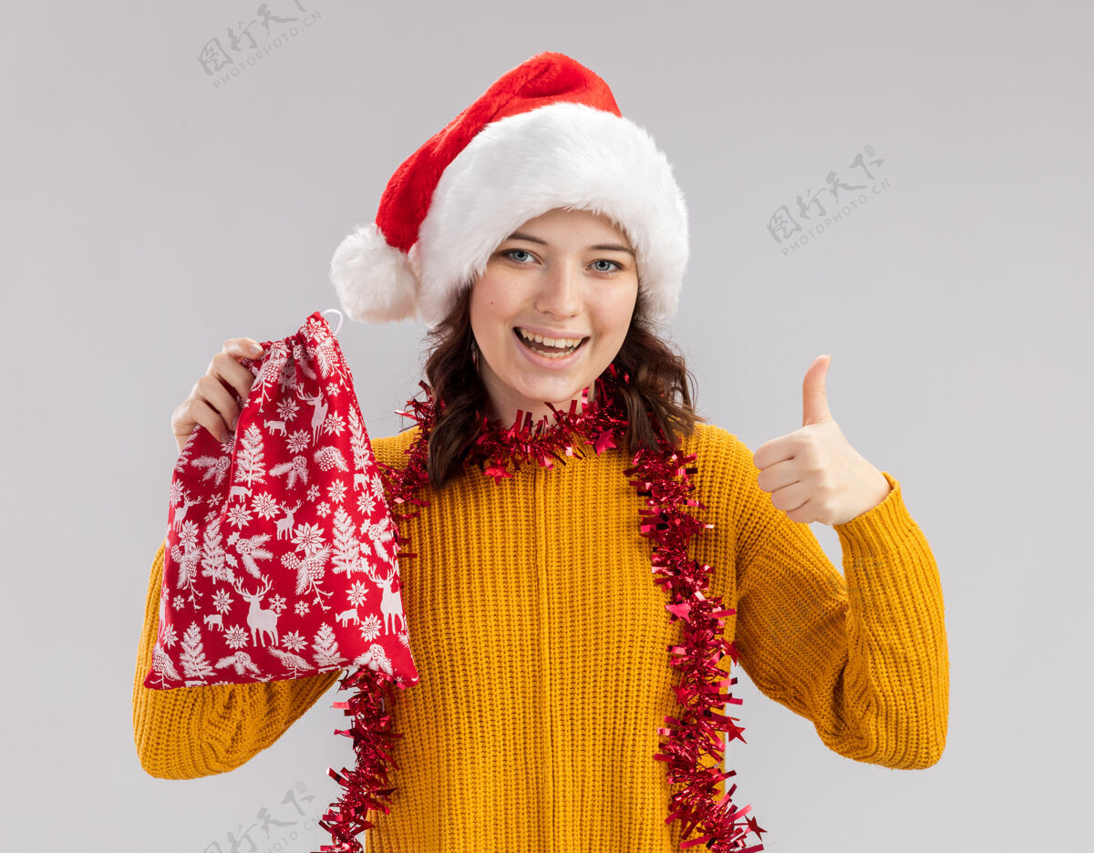 斯拉夫快乐的年轻斯拉夫女孩戴着圣诞帽 脖子上戴着花环 手里拿着圣诞礼品袋 竖起大拇指 与白色背景隔离 留有复制空间拇指举行礼物