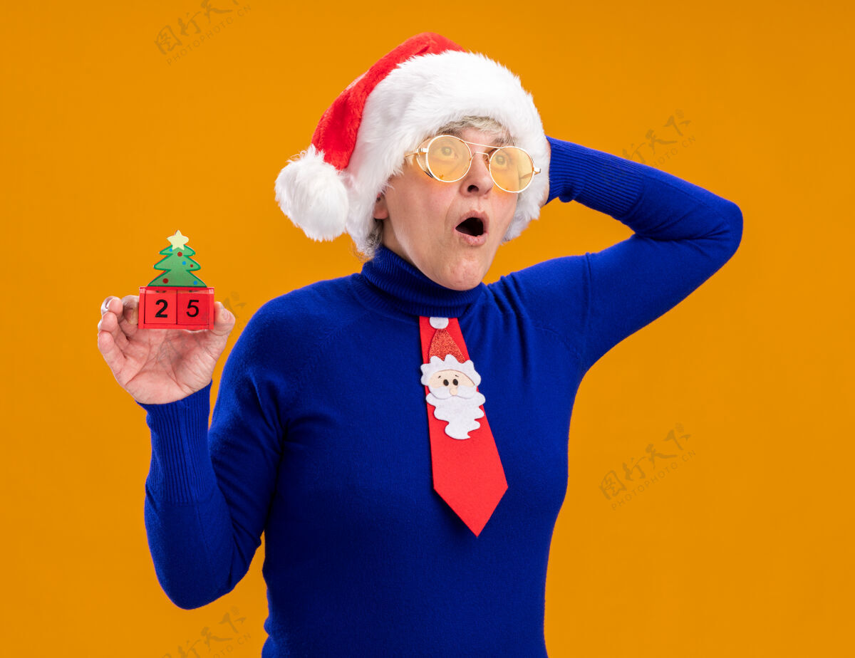 圣诞老人戴着太阳眼镜 戴着圣诞帽 系着圣诞领带的惊讶的老妇人拿着圣诞树饰物 把手放在头上 看着橙色背景上孤立的一面 还有复制空间惊喜帽子装饰品