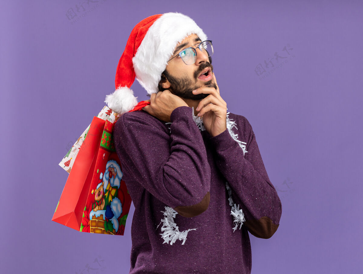 肩膀年轻帅哥戴着圣诞帽 脖子上戴着花环 肩上挎着礼品袋 手放在脸颊上 蓝色的背景上与世隔绝脸颊花环手