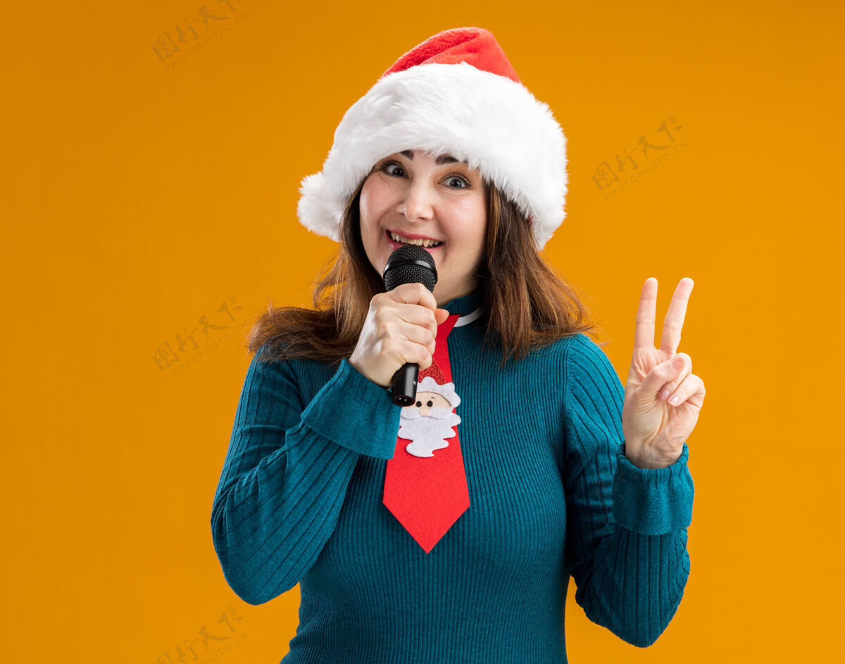 胜利微笑的成年白人妇女 戴着圣诞帽 打着圣诞领带 拿着麦克风 打着胜利的手势 橙色背景上有复制空间手势快乐麦克风
