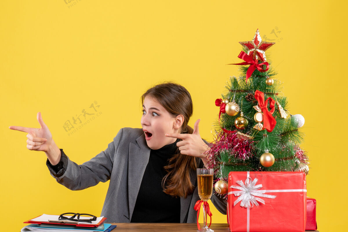 圣诞节正面图坐在桌边的大眼睛女孩用手指指着圣诞树和礼物鸡尾酒指着坐着公司