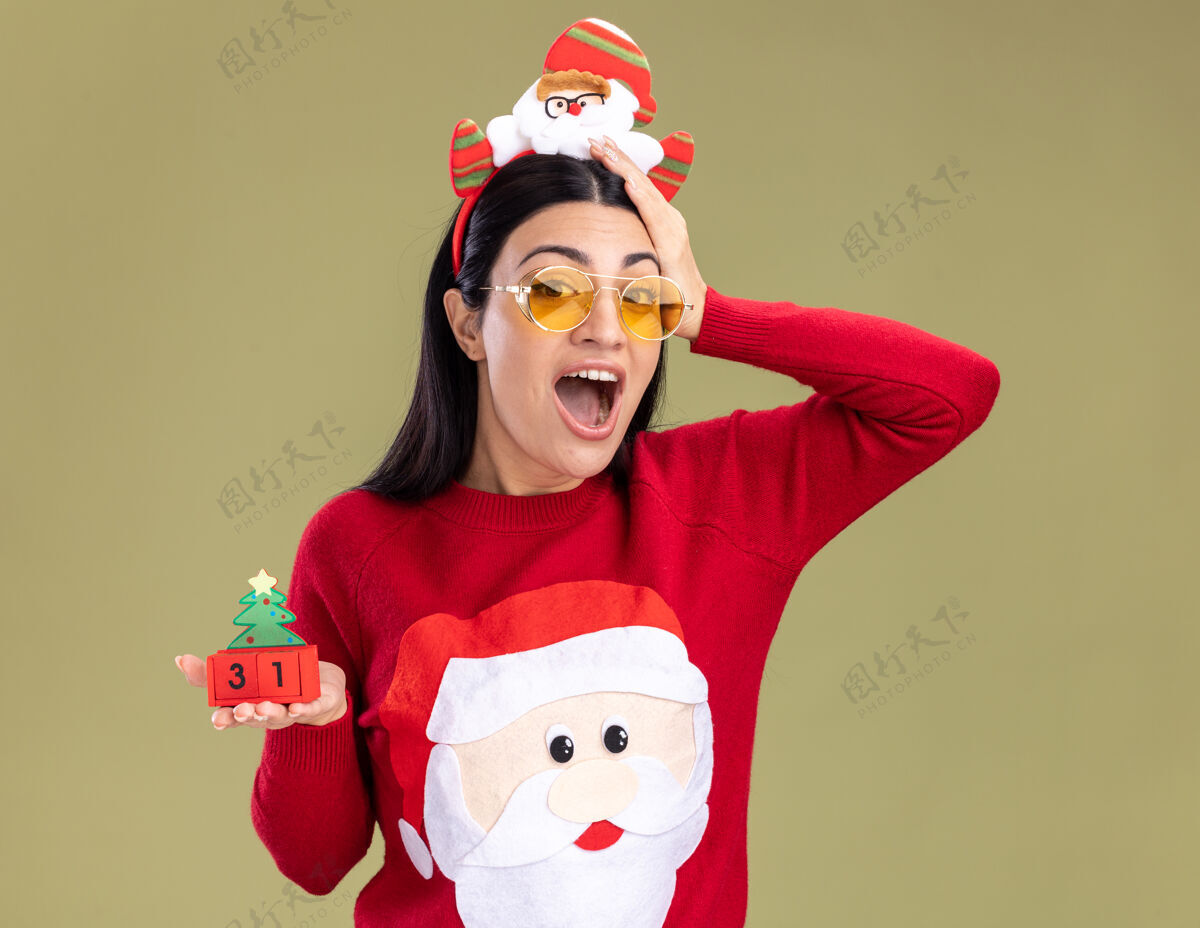 圣诞老人令人印象深刻的年轻白人女孩戴着圣诞老人的头带和带眼镜的毛衣拿着圣诞树玩具和日期保持手在头上隔离在橄榄绿的墙上日期橄榄毛衣