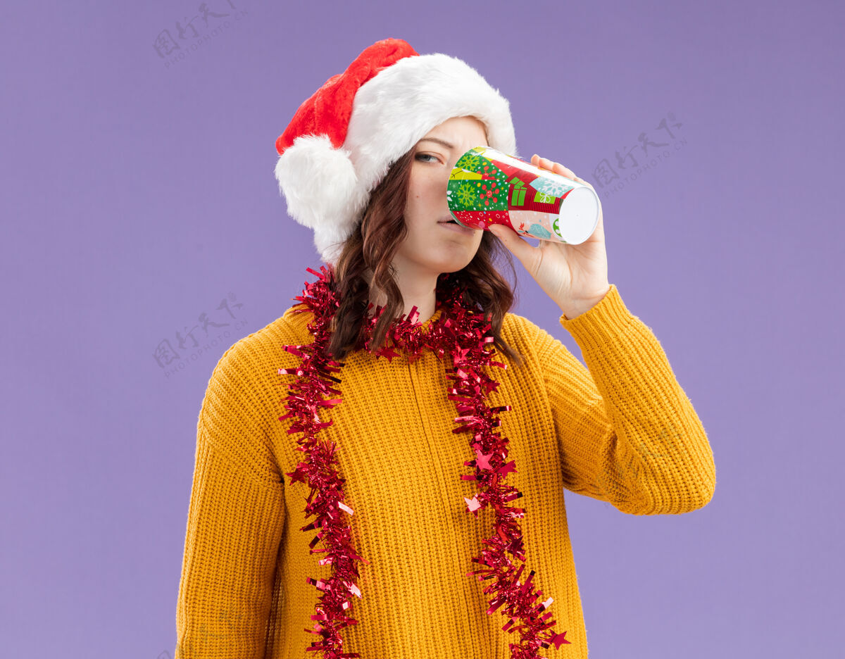 年轻自信的年轻斯拉夫女孩戴着圣诞帽 脖子上戴着花环 喝着纸杯里的酒 背景是紫色的 有复印空间周围花环斯拉夫人