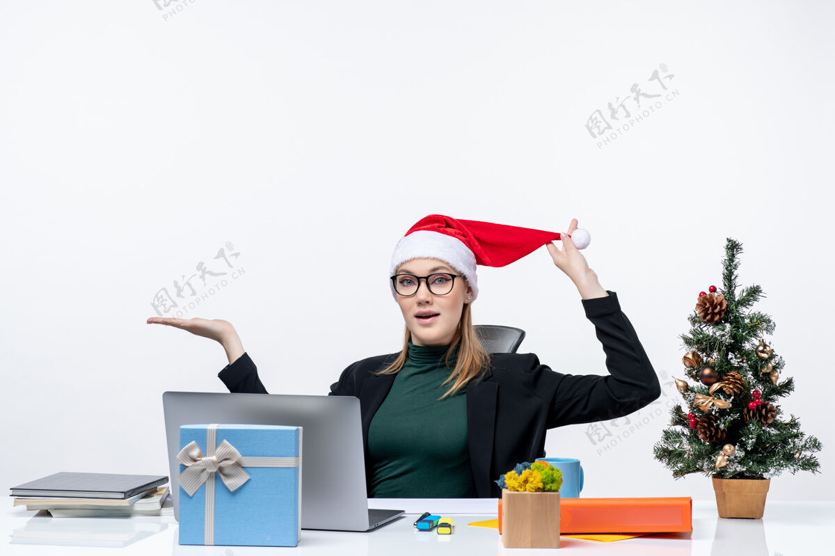 快乐一位年轻的女士拿着圣诞树和礼物坐在桌子旁 手里拿着圣诞老人的帽子和眼镜年轻女人坐着圣诞老人
