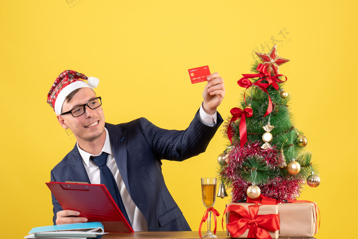 坐着一个商人坐在圣诞树旁的桌子旁看着他的卡片 黄色的礼物黄色商人树