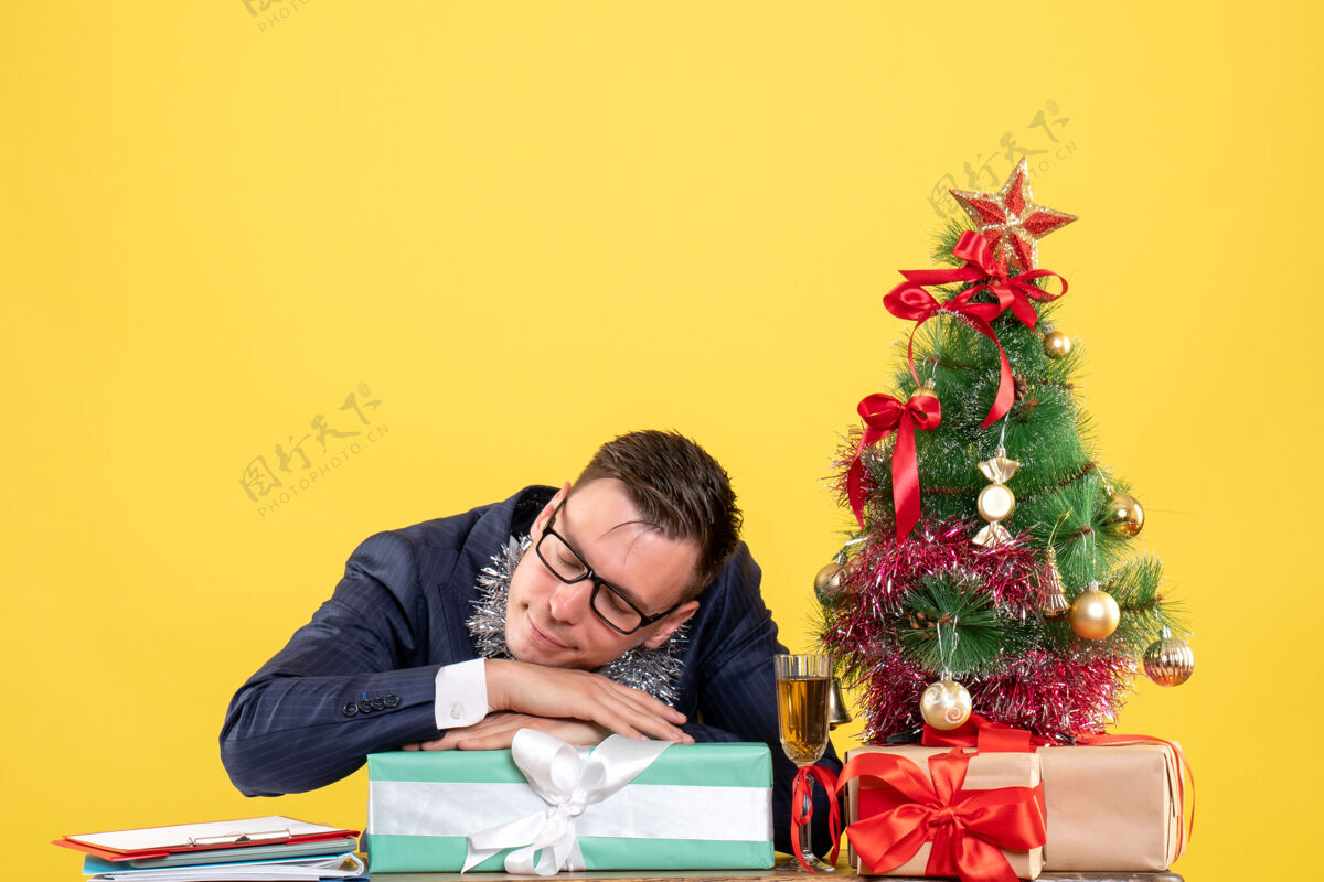 礼物昏昏欲睡的商人坐在圣诞树旁的桌子前 黄色的礼物坐着桌子新郎